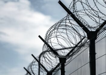 A prison border fence with razor wire against dark sky.
