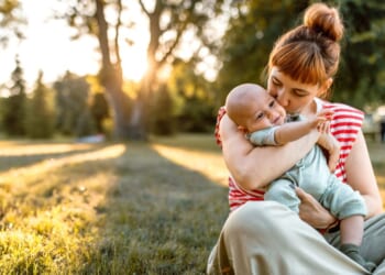 A mother and her baby embrace while sitting down in a meadow in sunset.