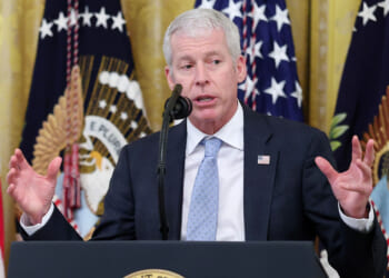 Energy Secretary Chris Wright holds his hands up while speaking behind a podium with flags behind him.