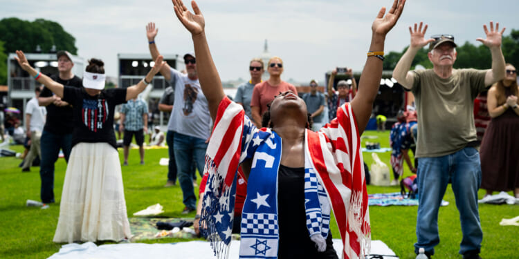 A Black woman draped in American flag and Israeli scarf lifts hands to heaven, with US Capitol in distant background.