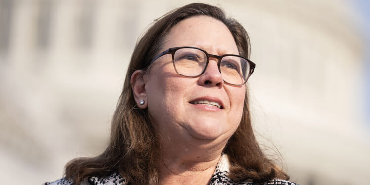 A woman with long brown hair, glasses, and a light-colored jacket stands outdoors in front of a blurred, domed building, looking slightly to the side and smiling.