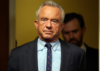 A man in a suit and tie stands indoors with a serious expression, facing forward. There is another person blurred in the background and the setting appears to be an office or hallway.