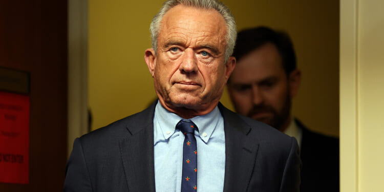 A man in a suit and tie stands indoors with a serious expression, facing forward. There is another person blurred in the background and the setting appears to be an office or hallway.