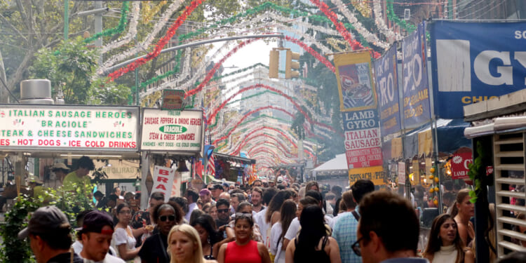 A crowded Italian street festival with food stalls, colorful hanging decorations, and many people walking and enjoying the event on a sunny day.