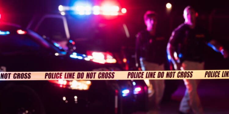 Two police officers standing in front of patrol cars, behind police tape, wearing bulletproof vests and duty belts.