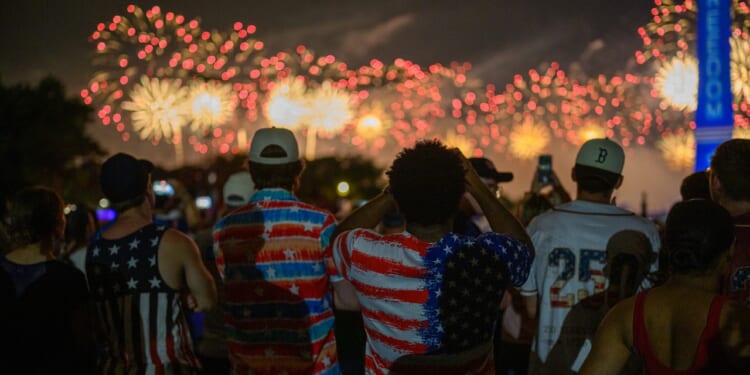 People dressed in American flag-themed clothing watch colorful fireworks light up the night sky, celebrating together outdoors.