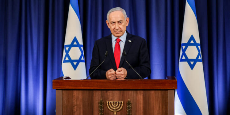 Benjamin Netanyahu stands at a podium with the Israeli emblem, flanked by two Israeli flags, delivering a speech about Israel in front of a blue curtain.