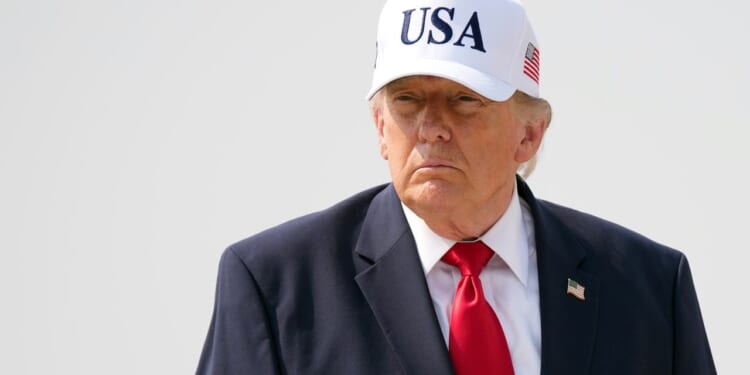 President Donald Trump wears a dark suit, red tie, and a white USA hat standing outside against a plain, light background.