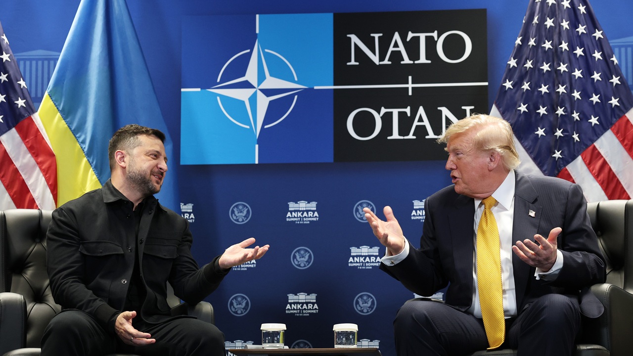 Two men in suits sit facing each other, gesturing while talking. Behind them are Ukrainian, NATO, and U.S. flags, along with a NATO OTAN logo.
