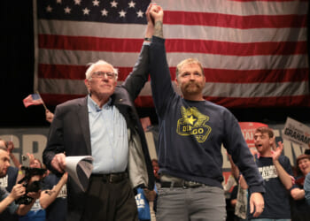 An older man in a suit holds up a mans arm on stage in front of a large American flag, with a cheering crowd in the background holding signs and waving flags.