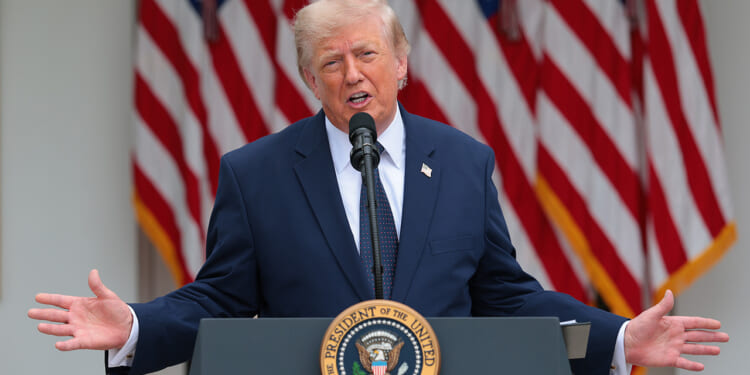 A man in a dark suit speaks at a podium with the U.S. presidential seal, gesturing with open arms. American flags are displayed in the background.