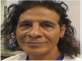 A person with long, dark curly hair and a slight smile faces the camera. They are wearing a light-colored shirt and are indoors, with a blurred background.
