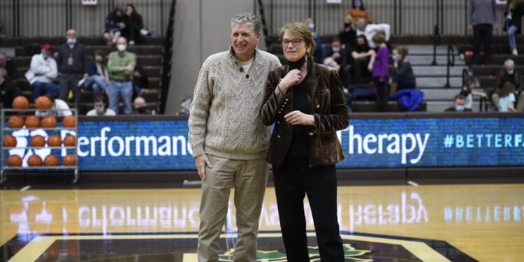 A man and a woman stand together on a basketball court, smiling. People are seated in the bleachers behind them, and a digital scoreboard displays the words Performance Therapy..