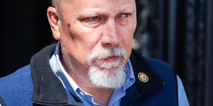 A bald man with a white goatee and a serious expression wears a blue vest and light blue shirt, standing outdoors with a dark background.