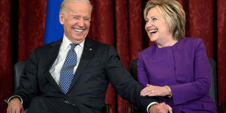 Two people in business attire sit side by side, smiling and laughing. The person on the left wears a dark suit and patterned tie; the person on the right wears a purple outfit. They are holding hands and appear to be enjoying a light moment.