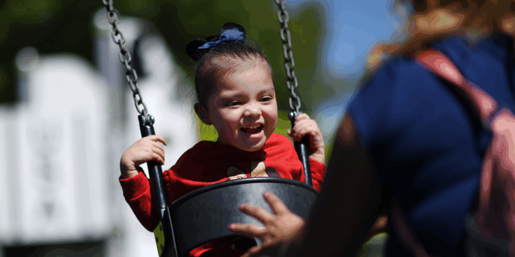 A young child smiles joyfully while swinging at a playground, wearing a red shirt and dark bow. An adult with a pink backpack stands nearby, gently pushing the swing. Trees and blurred playground equipment are in the background.