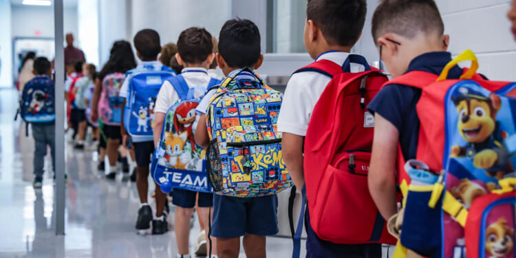 A group of young children, wearing backpacks and school uniforms, walk in a line down a brightly lit school hallway. The backpacks have colorful cartoon designs.