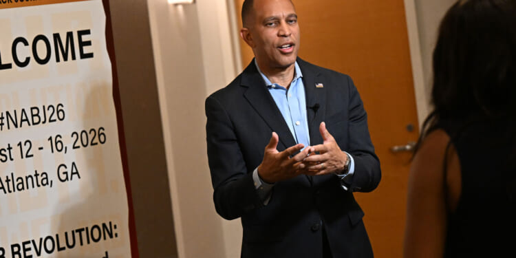 Hakeem Jeffries in suit speaks to a woman in front of a NABJ conference welcome sign in a hallway.