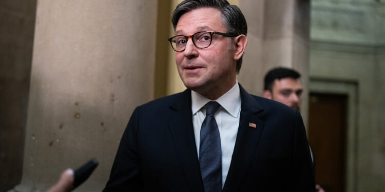 A man in a dark suit and tie, wearing glasses, stands indoors near large columns, speaking while being photographed or interviewed.