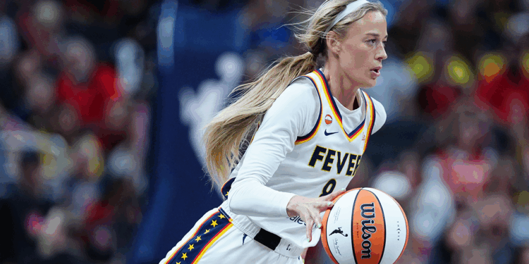 A basketball player in a white Fever uniform dribbles a basketball during a game, with focus and intensity, as the crowd watches in the blurred background.