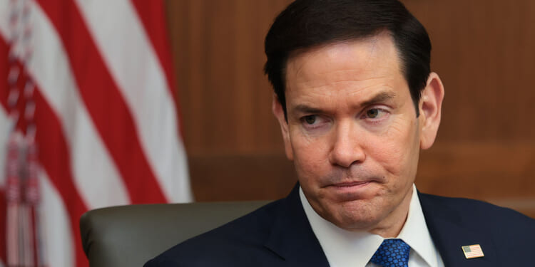 Secretary of State Marco Rubio designated the A/I Collective a terrorist group, noting its support for Jane's Revenge and other leftist terrorism. Pictured: Rubio, a man in a suit and tie sits in front of an American flag, looking to the side with a serious expression. An American flag pin is visible on his lapel.