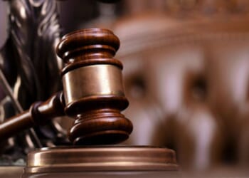 A gavel lined with gold sits on a wooden block ahead of a leather chair.