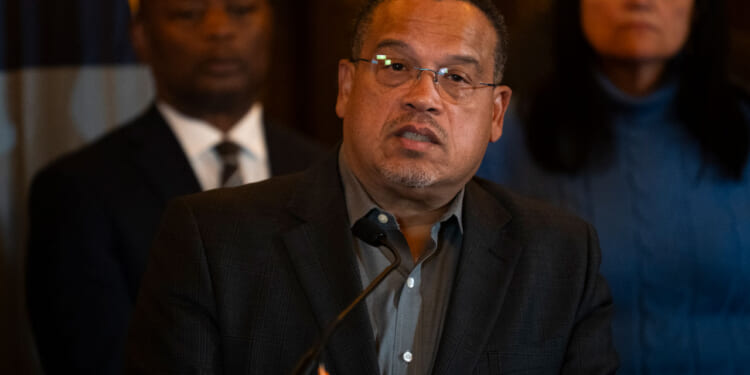 Keith Ellison glasses and a dark blazer speaks at a podium, with two people standing behind him, during an event in Minnesota.