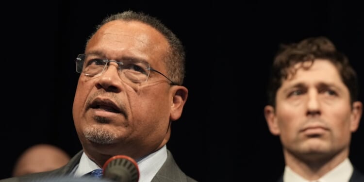 A man wearing glasses and a suit speaks at a microphone, with another man in a suit standing slightly behind him against a dark background.