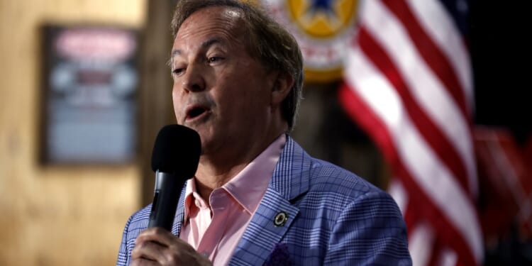 A man in a blue checkered blazer and pink shirt speaks into a microphone. An American flag and blurred background with signage are visible behind him.