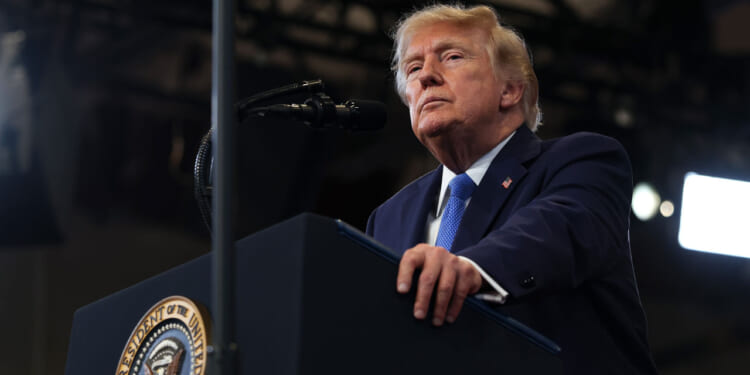 President Donald J. Trump wears a dark suit and blue tie stands at a podium with a presidential seal, speaking into a microphone in a well-lit room.