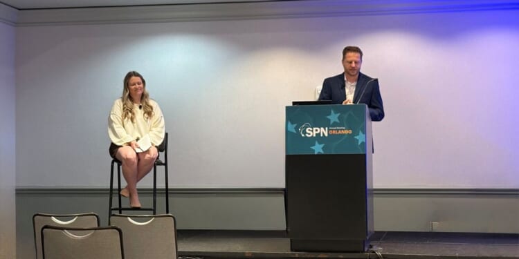 Camille Walsh, director of media relations at State Policy Network, sits on a tall chair, and Corey Burres, senior director of communication strategies at State Policy Network, stands at a podium labeled SPN Orlando in a conference room, with empty chairs in the foreground.