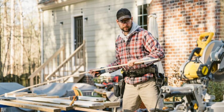 A man works in the yard of a house with wood and tools.