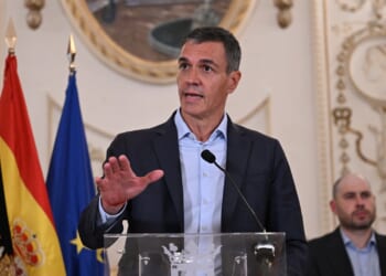 A man in a suit speaks at a podium with flags behind him, including the Spanish and EU flags, inside an ornate room. Another man is seated in the background, and a decorative round frame hangs on the wall.