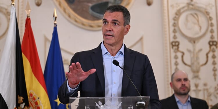 A man in a suit speaks at a podium with flags behind him, including the Spanish and EU flags, inside an ornate room. Another man is seated in the background, and a decorative round frame hangs on the wall.