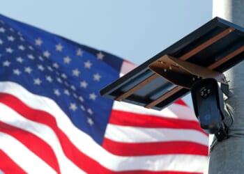 A Flock Safety camera is shown mounted in front of an American flag.
