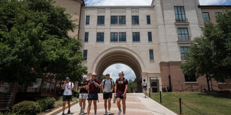 Six people walk on a pathway outside a large, beige university building with an archway, surrounded by trees under a partly cloudy sky.