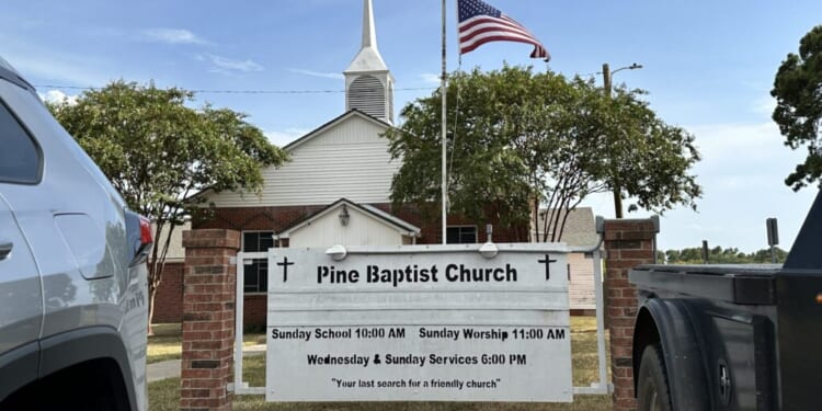 White church with a steeple and American flag, seen behind a sign reading Pine Baptist Church with service times, flanked by parked vehicles on grass. Trees frame the church against a blue sky.