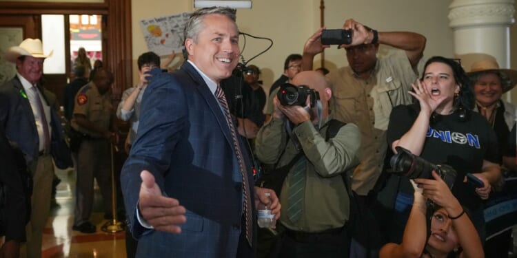 A man in a suit smiles and gestures while surrounded by photographers and people, some with cameras and others reacting energetically, at an indoor event.