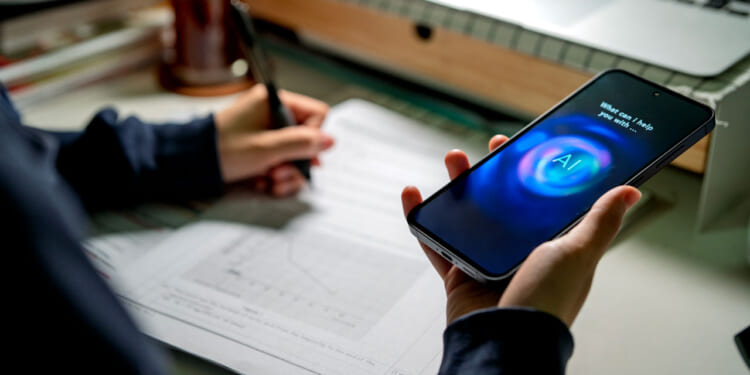 A person writes on paper while holding a smartphone displaying a digital assistant with What can I help you with? and AI on the screen, illustrating how technology reduces friction in daily tasks. A laptop and office supplies are on the desk in the background.
