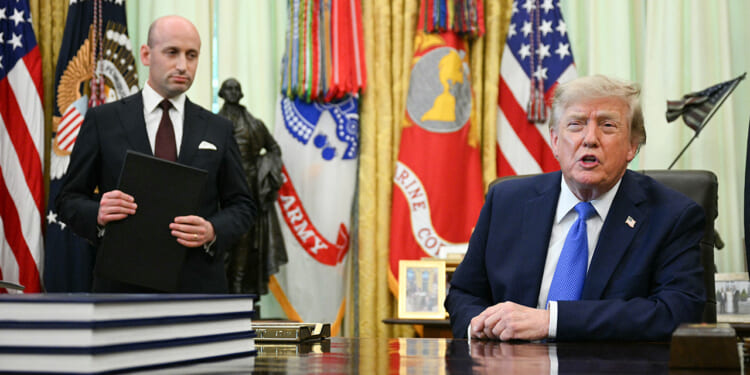 A man in a suit stands holding a folder while another man sits at a large desk, both in a room with military flags, the U.S. flag, and a gold curtain in the background.
