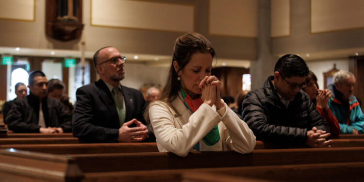 A group of people sits in church pews, heads bowed and hands clasped in prayer, in a softly lit sanctuary.