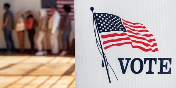 A voting booth stands in an auditorium with a line of citizens lined up behind it.