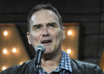 A man wearing a black jacket and checkered shirt speaks into a microphone, standing in front of a background with large, illuminated letters.