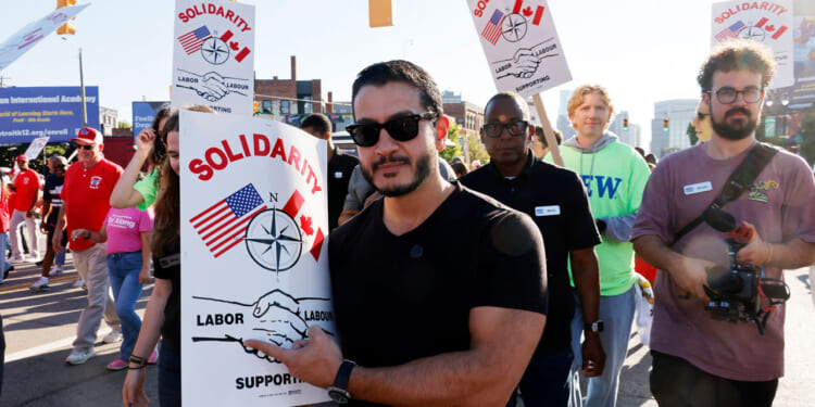 A group of people march on a street holding Solidarity signs with US and Canadian flags, a compass, and shaking hands, supporting labor unions and advocating for fair election practices. Abdul El-Sayed in sunglasses holds a sign in the front.
