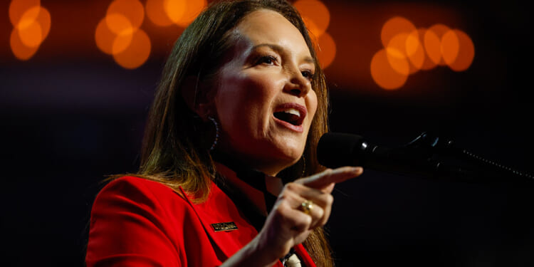 Brooke Rollins in a red blazer speaks passionately into a microphone, gesturing with her hand. Warm orange lights create a soft, out-of-focus background.