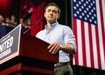 Sen. Jon Ossoff stands at a podium labeled UNITED FOR GEORGIA with an American flag in the background and people watching from the side.