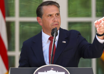 A man in a dark suit and red tie speaks at a podium outdoors, gesturing with his right hand as he addresses concerns about election fraud. American flags and green foliage are in the background.