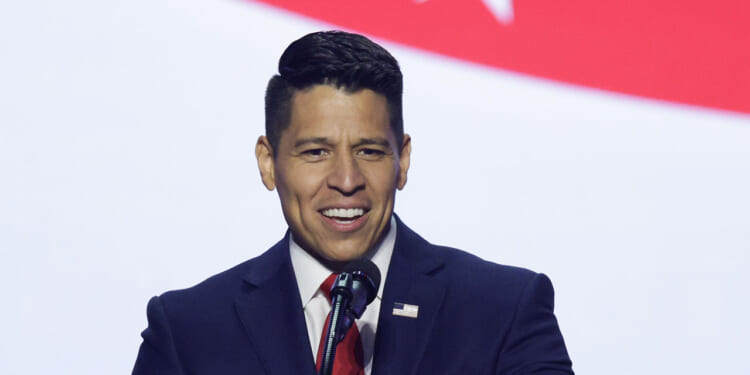 A man in a dark suit and red tie speaks at a podium with a microphone, against a backdrop featuring part of a red stripe and a white star, representing the Texas 35th Congressional District.