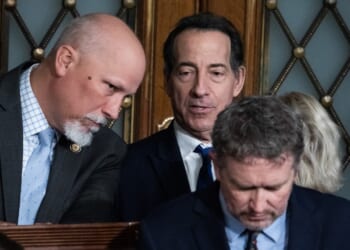 Three men in suits are gathered closely together, appearing to have a serious discussion. Two men are standing, one is leaning in to speak, while the third man sits in front of them, looking down. Ornate wooden doors are in the background.