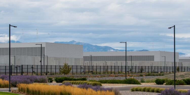 A large, modern industrial data center building with gray walls and flat roofs. It sits behind a black fence, surrounded by landscaping with grasses and shrubs, with mountains and cloudy skies in the background.